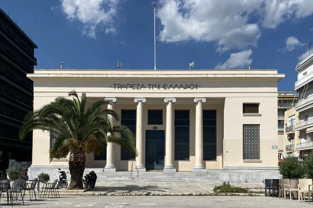 Φωτογραφία με νεοκλασικό κτίριο τράπεζας με κίονες και φοίνικα στην είσοδο, με ανοιχτό ουρανό