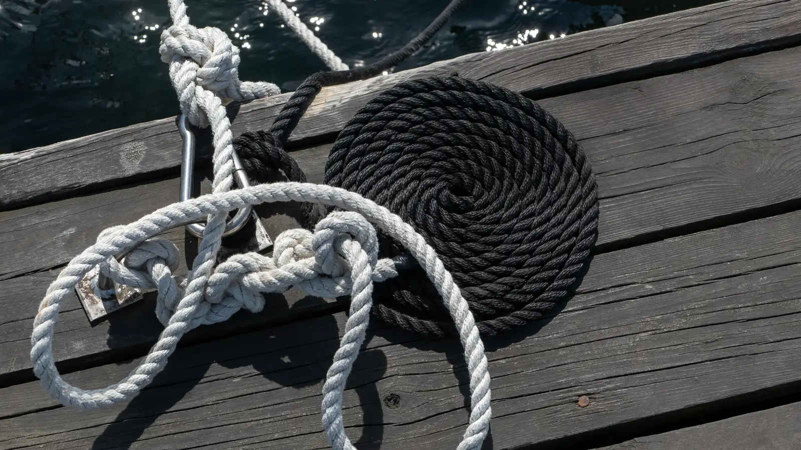 Φωτογραφία με λευκό σχοινί δεμένο σε κόμπους και μαύρο κουλουριασμένο σχοινί πάνω σε ξύλινη προβλήτα